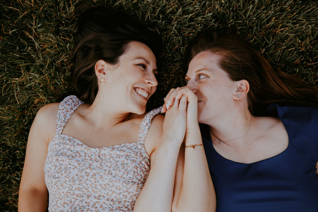 An LGBTQ+ couple laying in the grass, looking at each other, holding hands. 