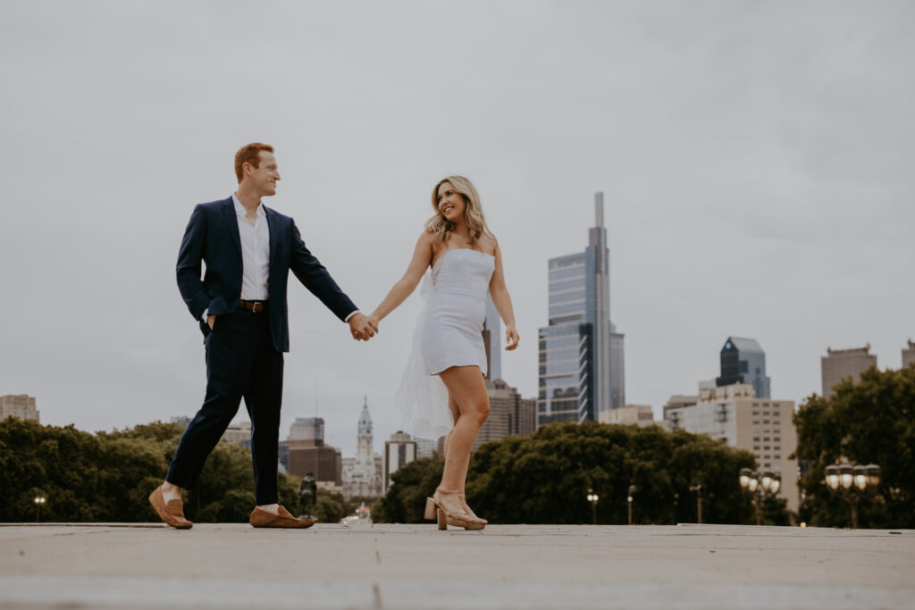 A couple holding hands, walking, with the Philadelphia skyline behind them. 