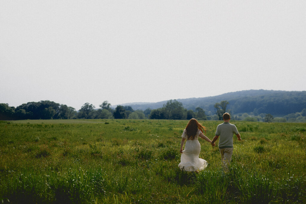 A couple running through an open field, holding hands. 