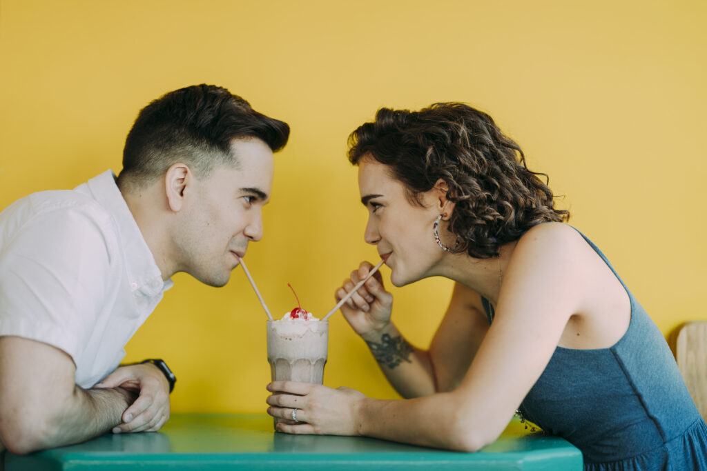 A couple drinking from a shared mikshake at a green table against a yellow wall. 