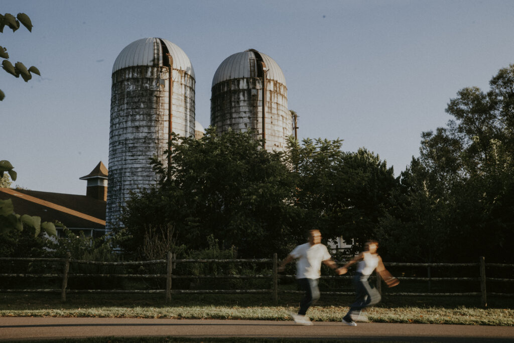 Two people holding hands, running, with farm silos in the background. 